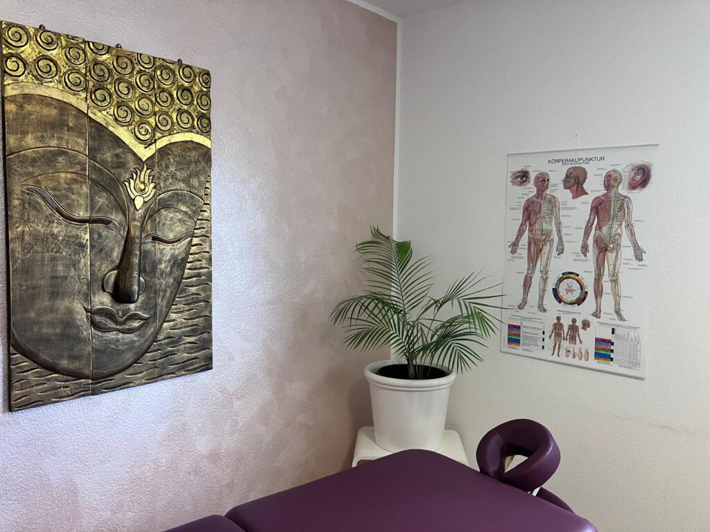 Behandlungsraum mit einem buddhistischen Bild und einem Poster des menschlichen Körpers. In der Ecke steht eine Grünpflanze.